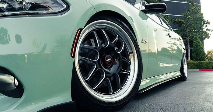 Close-up of a green car featuring WELD Racing Wheels on a smooth driveway with a modern building in the background