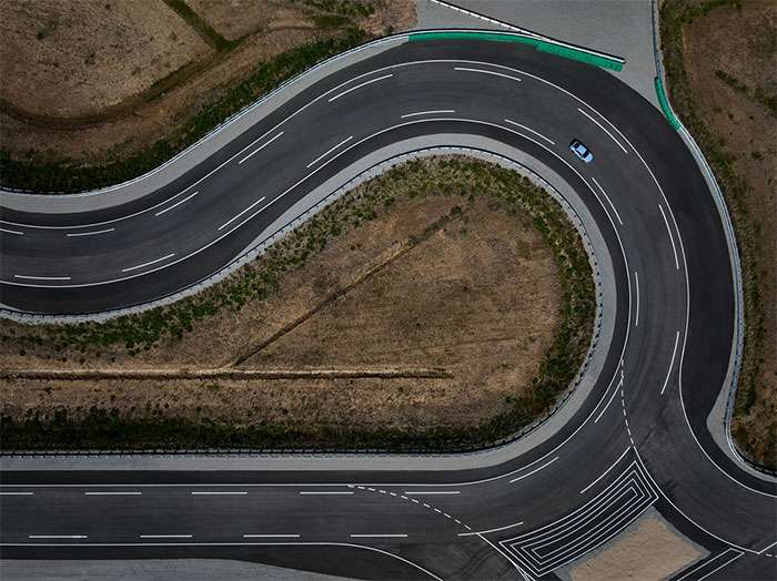 Aerial view of a blue car testing tires on a curved state-of-the-art asphalt test track surrounded by dry grass
