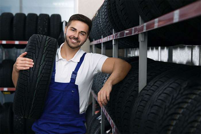 Mechanic in blue overalls holding a tire in a storage room filled with neatly stacked tires on metal racks.