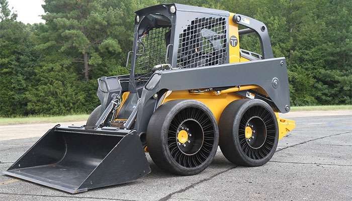 Skid-steer loader equipped with Michelin Tweel airless tires parked on pavement near trees.