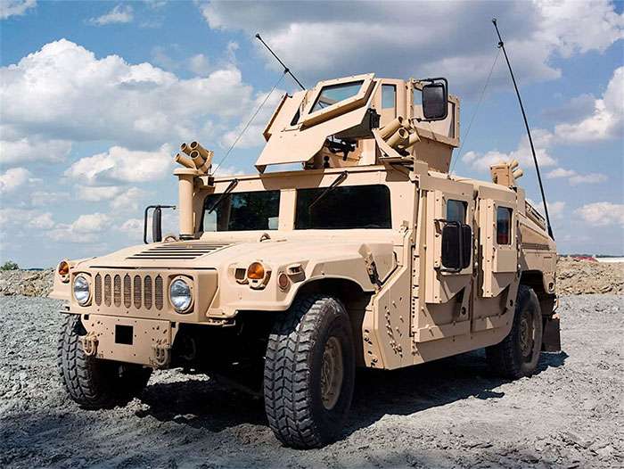 Tan military Humvee equipped with run-flat tires parked on rocky terrain under a partly cloudy sky