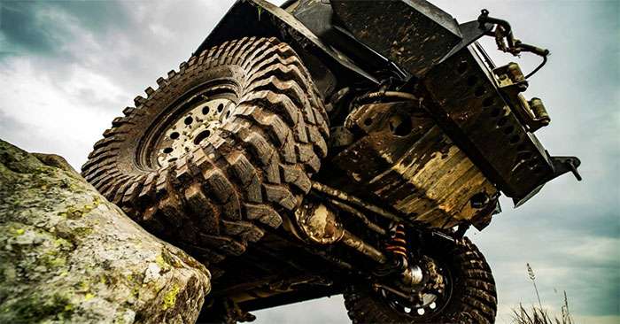 Close-up of muddy Nitto truck tires gripping rocky terrain under a rugged off-road vehicle against a cloudy sky