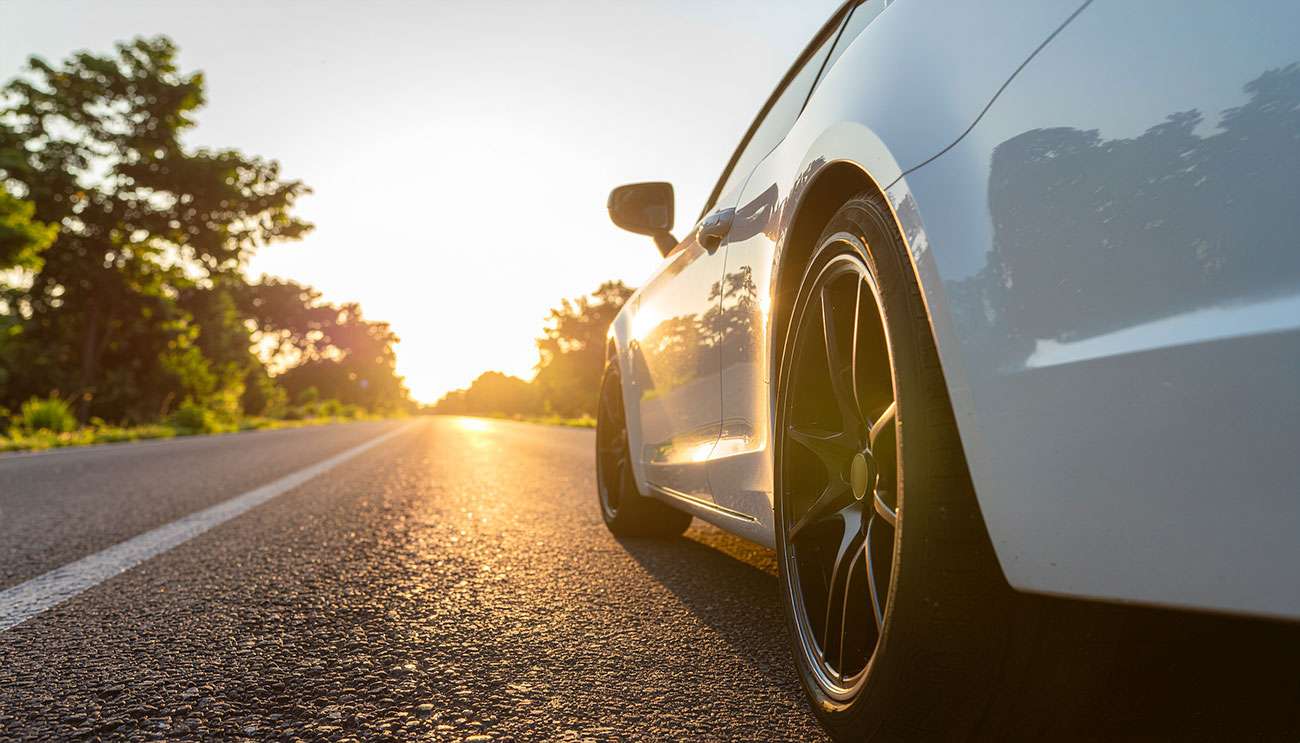 Close-up of a muscle car's rear tire and wheel on a sunlit road at sunset, highlighting tire tread and rim details