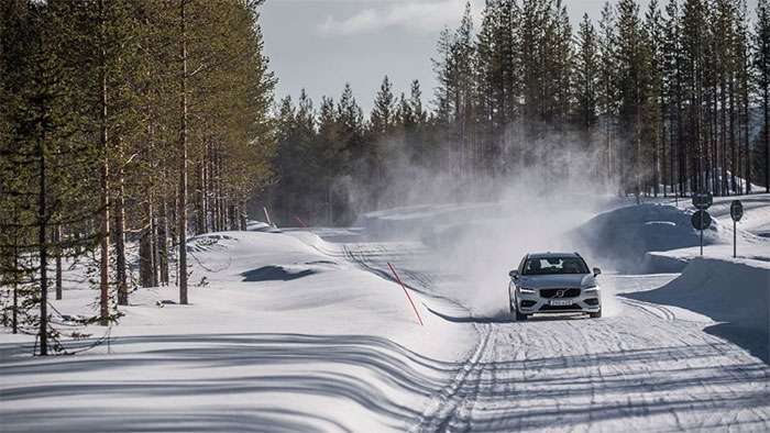 A car drives on a snowy road kicking up snow during the 2019 NAF Winter Tire Test in a forested area.