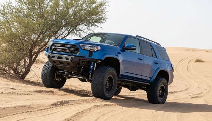 Blue off-road SUV with upgraded suspension and large tires parked on sandy terrain under a tree near a desert road
