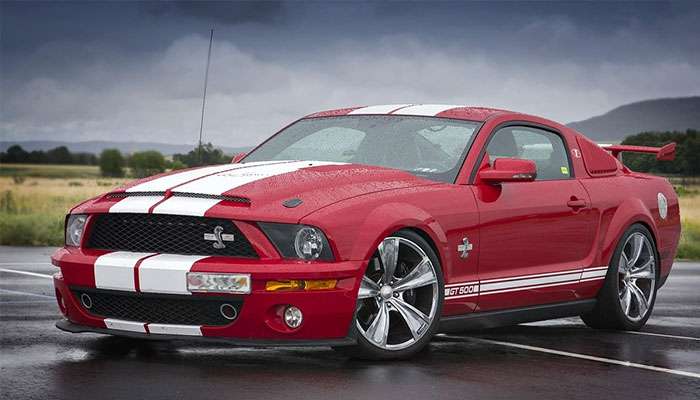 Red Ford Mustang Shelby GT500 muscle car with white racing stripes parked on wet pavement under a cloudy sky