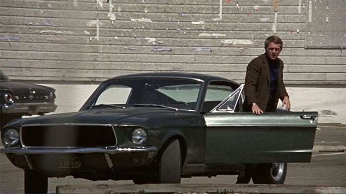 Steve McQueen standing beside a classic dark green 1968 Ford Mustang from the movie Bullitt on a city street.