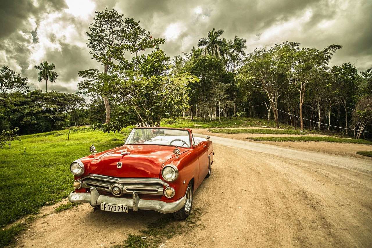 Red vintage convertible car parked on a dirt road surrounded by lush greenery under a cloudy sky in Cuba.