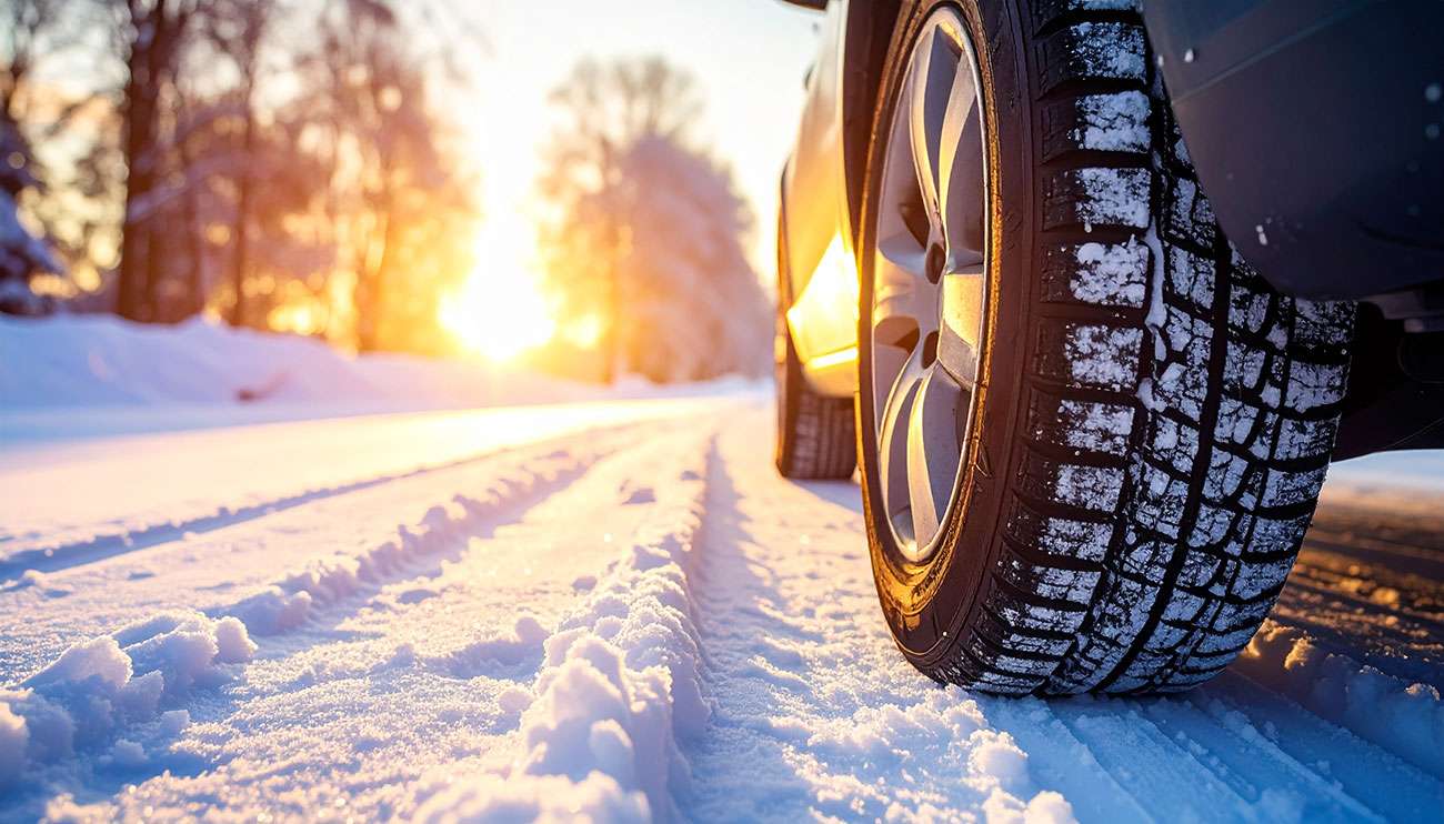 Close-up of a car tire with snow on the tread, parked on a snowy and icy road at sunset