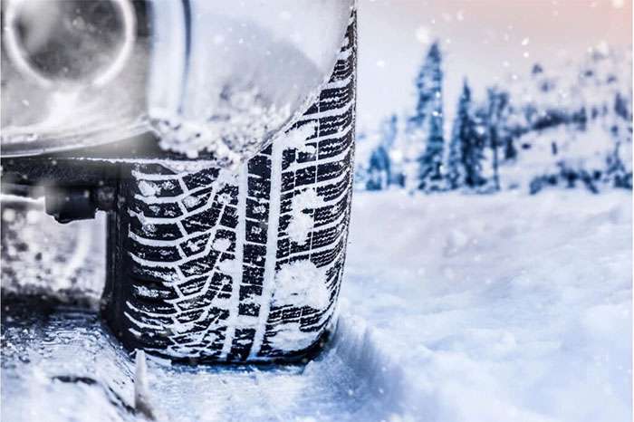 Winter tire gripping snow-covered ground with falling snow and evergreen trees in the background, close-up tread contact